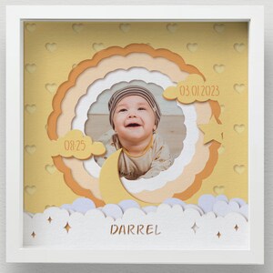 May include: A framed baby photo with a layered paper design. The design includes a smiling baby, a crescent moon, clouds, stars, and the name "DARREL". Dates and times are also included in the design. The frame is white.