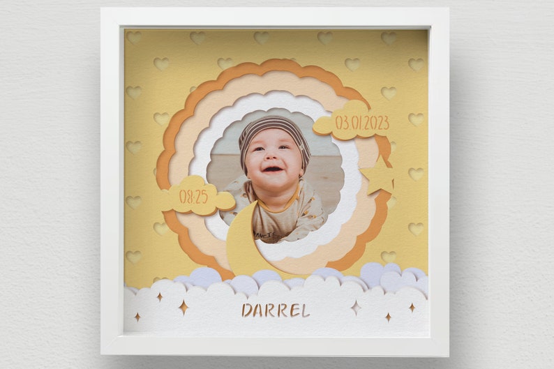 May include: A framed baby photo with a layered paper design. The design includes a smiling baby, a crescent moon, clouds, stars, and the name "DARREL". Dates and times are also included in the design. The frame is white.