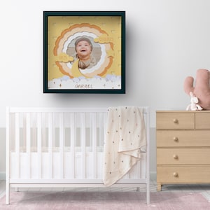 May include: A framed nursery art piece featuring a baby's photo within a layered, cloud-themed design. The frame is dark green, and the artwork includes the baby's name, "Darrel", and the date "05/05/2023". The nursery setting includes a white crib, a dresser, and a cactus-shaped pillow.