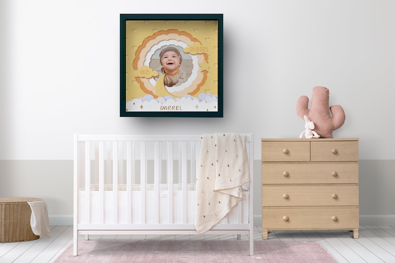 May include: A framed nursery art piece featuring a baby's photo within a layered, cloud-themed design. The frame is dark green, and the artwork includes the baby's name, "Darrel", and the date "05/05/2023". The nursery setting includes a white crib, a dresser, and a cactus-shaped pillow.