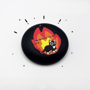 May include: Black round stone with a black cat on fire with the text "I do believe I'm on fire" printed on it.