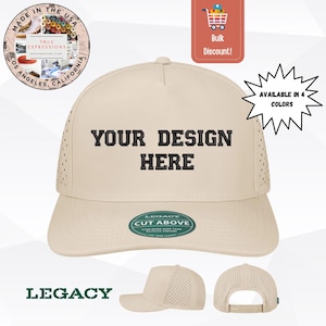 May include: A beige baseball cap featuring a five-panel construction with laser-cut ventilation holes on the side panels. The front panel displays the black embroidered text YOUR DESIGN HERE. A circular sticker is on the brim with the text LEGACY CUT ABOVE.