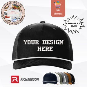 May include: Black trucker hat with white text that reads "YOUR DESIGN HERE". The hat has a white rope detail and a mesh back. The image also shows the Richardson logo and a color selection of seven different hats.