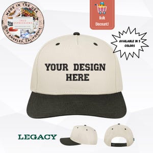 May include: A two-tone baseball cap with a cream-colored crown and a black brim. The front of the crown features the black embroidered text "YOUR DESIGN HERE". The cap includes a black button on top and a black adjustable snapback closure at the rear.