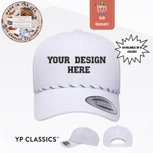 May include: A white baseball cap featuring black embroidered text that reads YOUR DESIGN HERE on the front panel. The cap includes a decorative white and black twisted rope detail across the base of the crown and a YP Classics sticker on the brim.