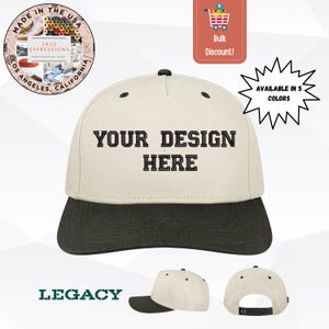 May include: A two-tone baseball cap with a cream-coloured crown and a dark green brim. The text "YOUR DESIGN HERE" is embroidered in black. The cap is part of the Legacy collection and is available in 5 colours.