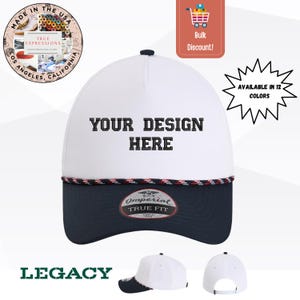 Könnte beinhalten: Eine weiße Baseballkappe mit dunkelblauem Schirm und einer dekorativen Kordelverzierung an der Basis der Krone. Auf der Vorderseite befindet sich der schwarze Schriftzug YOUR DESIGN HERE. Ein Imperial True Fit Aufkleber ist auf dem Schirm.
