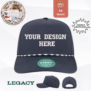 Puede incluir: Gorra azul marino con texto blanco bordado "YOUR DESIGN HERE". La gorra tiene un detalle de cuerda blanca y azul marino a lo largo del borde. La gorra está etiquetada como "LEGACY CADDY" y está disponible en varios colores.