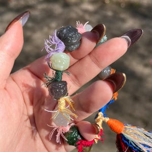 Pode incluir: Um bracelete colorido com contas de jade verde, branco e preto, e um tassel multicolorido. O bracelete é feito com uma variedade de materiais, incluindo tecido, corda e contas.