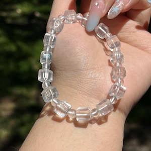 May include: A clear quartz bracelet with faceted, cube-shaped beads. The translucent beads are strung together. The bracelet is held in a hand, highlighting its design and clarity. The bracelet is a piece of jewellery.