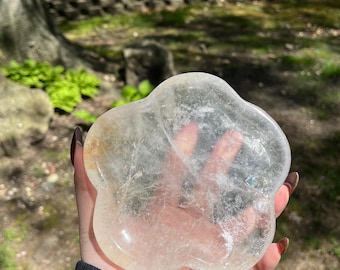 May include: A clear quartz crystal carved into a flower shape. The crystal is translucent and has a slight yellow tint. It is being held in a hand.