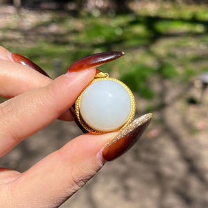 May include: A white jade pendant with a gold rim, held in a hand with brown nail polish.