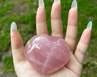Peut inclure: Un cristal de quartz rose en forme de cœur, tenu dans une main ouverte. Le cristal est d'une couleur rose tendre et a une surface lisse et polie. L'arrière-plan est de l'herbe verte floue.