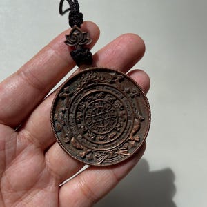 May include: A circular copper-toned pendant featuring intricate embossed zodiac symbols and detailed patterns arranged in concentric rings. The pendant is attached to a black braided cord with a small lotus-shaped bead accent at the top.