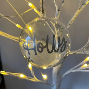 May include: A clear, spherical Christmas bauble with the name "Holly" in a cursive font. The bauble contains a snowy filling and a snowflake design. It hangs from a twine string and is lit by small, warm-toned fairy lights.
