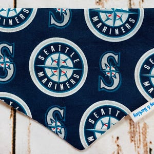 Può includere: Un bandana blu con una bussola bianca e le parole "Seattle Mariners" ripetute su tutto il tessuto. Il bandana ha un'etichetta bianca con il testo "Leroy & Barkley" su di essa.
