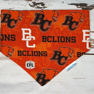 Op de afbeelding: Oranje en zwarte bandana voor huisdieren met de tekst "BC Lions" en een logo van een poema-kop. De bandana heeft ook het CFL-logo en een klein label met "Leroy & Barkley".