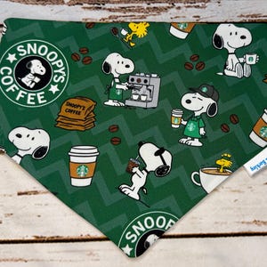 Peut inclure: Bandana verte pour chien avec un motif répété de personnages Snoopy, de tasses à café et de grains de café. La bandana porte le texte "Snoopy's Coffee" et un logo inspiré de Starbucks. Le fond présente un motif en chevrons.