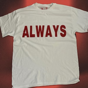 May include: A white short-sleeved t-shirt featuring the word ALWAYS printed in large, bold, dark red capital letters across the chest. The garment has a crew neckline and a small interior label visible at the collar. The text size is large.