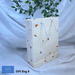 May include: A white gift bag with a white rope handle. The bag is made of textured paper with small orange and blue flower petals embedded in the paper. The bag is sitting on a white surface. "Gift Bag 8" is written in black text below the bag.