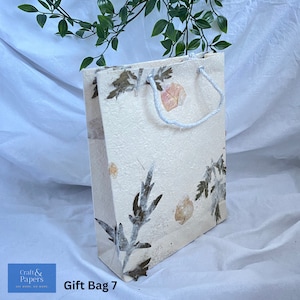 May include: A white gift bag with a floral design made from pressed flowers and leaves. The bag has a white rope handle and the text "Gift Bag 7" is printed on the bottom.