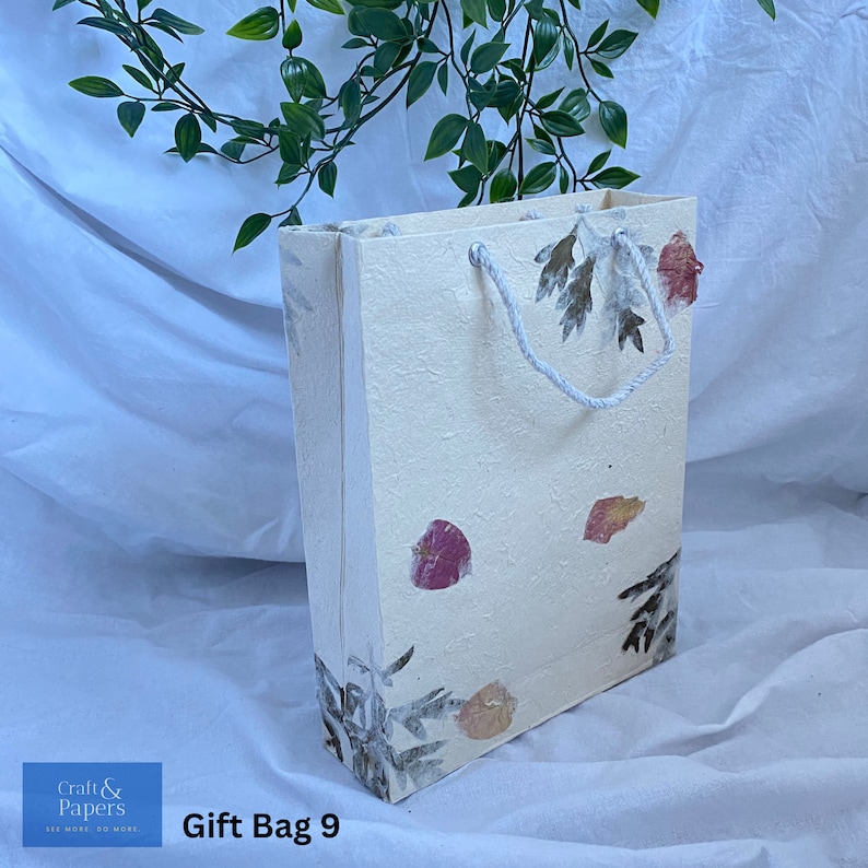 May include: A white gift bag with a floral design and a white rope handle. The bag has a textured surface and features pressed flowers and leaves. The text "Gift Bag 9" is printed on the bottom of the bag.