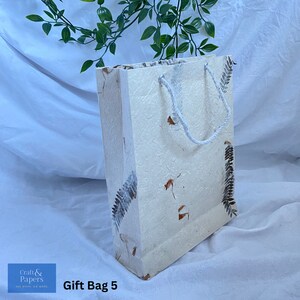 May include: A white gift bag with a white rope handle and brown leaf designs. The bag is made of textured paper and has the text "Gift Bag 5" printed on the bottom.