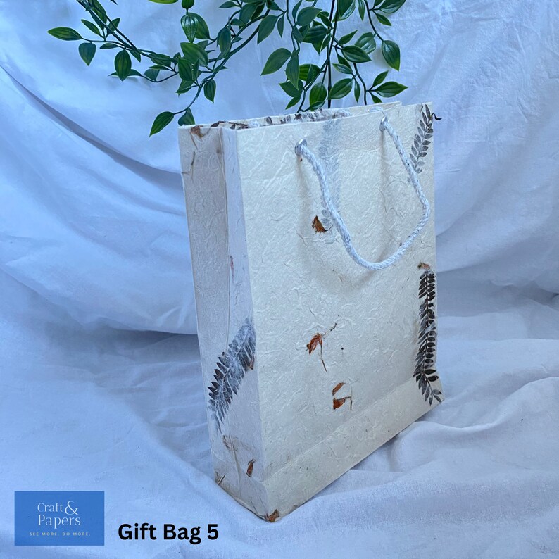 May include: A white gift bag with a white rope handle and brown leaf designs. The bag is made of textured paper and has the text "Gift Bag 5" printed on the bottom.