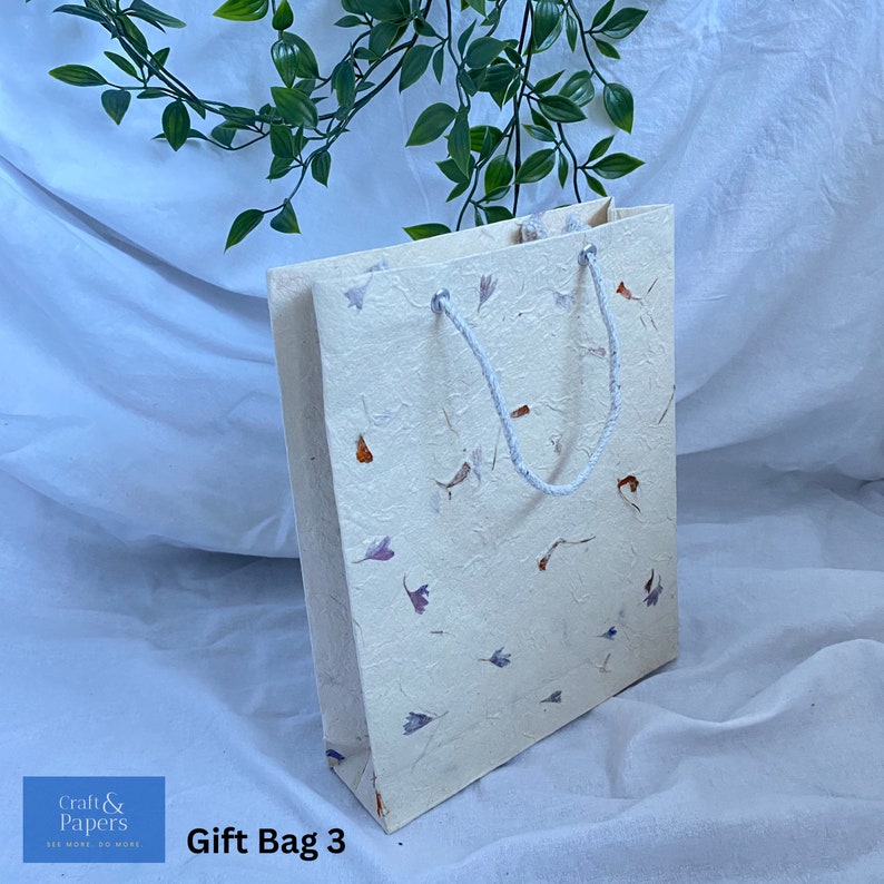 May include: A white gift bag with a floral pattern and a white rope handle. The bag is made of recycled paper and has the text "Craft & Papers" on the bottom left corner. The text "Gift Bag 3" is on the bottom right corner.