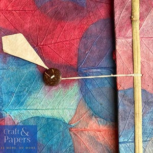 May include: A handmade journal with a colorful cover made of pressed leaves. The cover features a blue and red leaf pattern with a white string and a coconut shell button. The journal has the text "Craft & Papers" and "See More. Do More." printed on the front.