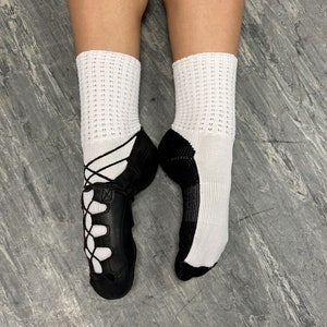 May include: A pair of black leather Irish dance shoes with white laces and white socks. The shoes have a decorative pattern on the front.