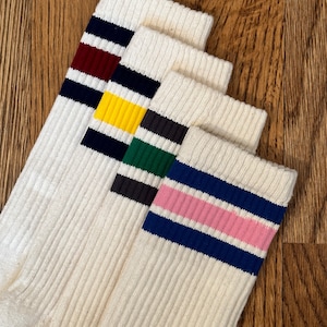 May include: Four pairs of off-white athletic socks with colored stripes. Each pair features a different stripe color: navy and burgundy, navy and yellow, black and green, and navy and pink. The socks are ribbed and made of a soft material.