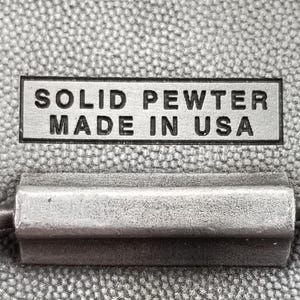 May include: Close-up of a rectangular, silver-colored metal tag with the words "SOLID PEWTER MADE IN USA" in black lettering. Below the tag is a silver-colored metal object. The background is a textured, gray fabric.