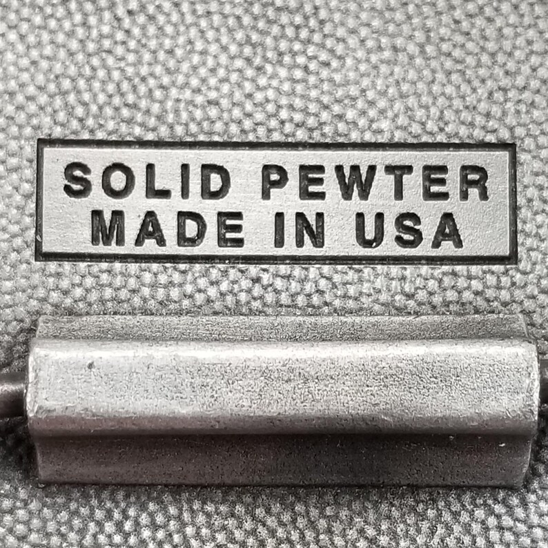 May include: Close-up of a rectangular, silver-colored metal tag with the words "SOLID PEWTER MADE IN USA" in black lettering. Below the tag is a silver-colored metal object. The background is a textured, gray fabric.