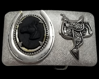 May include: Silver-toned rectangular belt buckle with a textured surface. Features a horseshoe and a black horse head cameo. A detailed saddle design is on the right side. The buckle has a western theme.