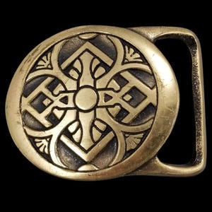 May include: A brass-colored belt buckle with an intricate, symmetrical design. The circular buckle features a central cross-like pattern surrounded by geometric shapes and floral accents. The buckle has a rectangular loop for the belt.