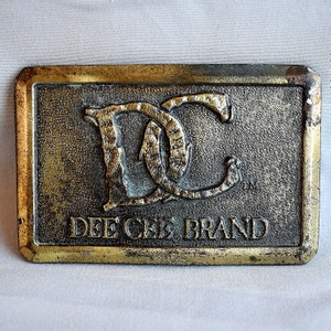 May include: Rectangular brass belt buckle with the words "DEE CEE BRAND" and the intertwined letters "DC". The buckle has a textured background and a gold-coloured border.