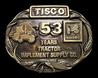 May include: A vintage brass belt buckle with the text "TISCO" at the top. The buckle features a tractor and the number "53" with the words "YEARS TRACTOR IMPLEMENT SUPPLY CO." The design includes wheat and corn.