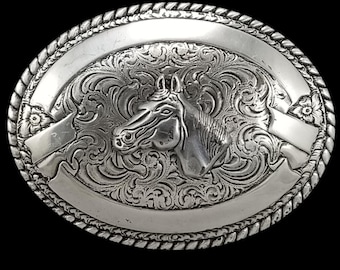 May include: Oval silver-toned belt buckle with a detailed horse head design. The buckle features intricate scrollwork and a rope-like border. The horse head is centered, surrounded by floral and banner elements.