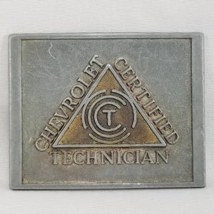 Könnte beinhalten: Eine silberne Metallplakette mit einem goldenen Dreieck in der Mitte. Das Dreieck enthält einen Kreis mit einem "T" darin und konzentrischen Kreisen darum herum. Der Text "CHEVROLET CERTIFIED TECHNICIAN" ist um das Dreieck herum geschrieben.