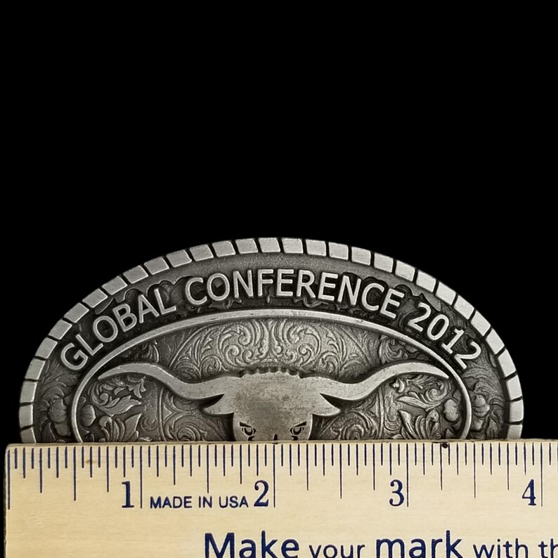 May include: A silver-colored metal belt buckle with the text "GLOBAL CONFERENCE 2012" and a bull's head design. The buckle is measured with a ruler, showing it is approximately 4 inches wide. The text "MADE IN USA" is also visible.