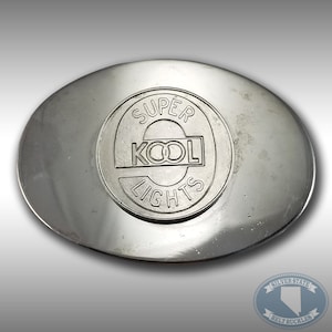 Può includere: Una fibbia per cintura in argento con un design circolare che presenta le parole "SUPER KOOL LIGHTS" in nero.