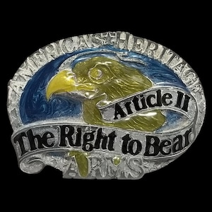 May include: A silver-colored belt buckle with an eagle design. The eagle is gold and green, set against a blue background. The buckle has the text "America's Heritage" and "Article II The Right to Bear ARMS".