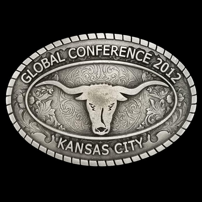 May include: Oval silver-toned belt buckle with a longhorn steer design. The buckle is engraved with the words "GLOBAL CONFERENCE 2012" and "KANSAS CITY". The background features a floral pattern.