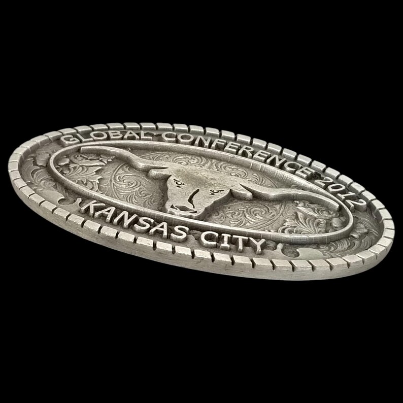 May include: Oval silver-colored belt buckle with the text "GLOBAL CONFERENCE 2012" and "KANSAS CITY". The buckle features a longhorn steer design in the center with decorative scrollwork.