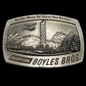 Könnte beinhalten: Silberfarbene Gürtelschnalle mit detaillierter Landschaftsszene. Die Schnalle zeigt eine Bohranlage, einen Hubschrauber, Berge und den Text "On The Move To Serve You Better" und "Norton Boyles Bros."