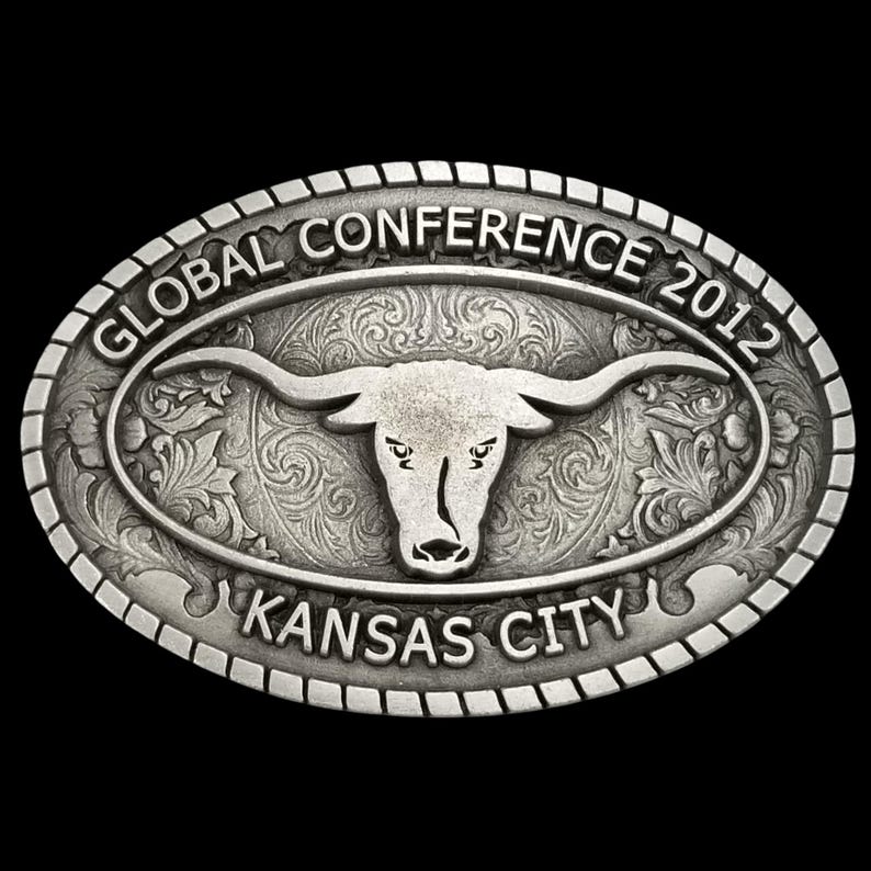 May include: Oval silver-colored belt buckle with a longhorn steer design. The buckle features the text "GLOBAL CONFERENCE 2012" and "KANSAS CITY" in a decorative font. The background has an ornate floral pattern.
