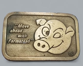 Op de afbeelding: Een gouden metalen gesp met een cartoon varkensgezicht en de tekst "Move ahead with Farmstead".