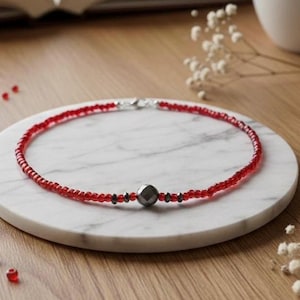 May include: A red beaded necklace with a silver clasp and a central dark gray geometric bead. The necklace is displayed on a white marble coaster, with a wooden surface in the background. Small red beads are scattered around.