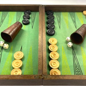 Op de afbeelding: Backgammon bordspel met een groene speeloppervlakte, houten frame en bruine leren dobbelbekers. Het spel bevat zwarte en beige schijven en dobbelstenen.